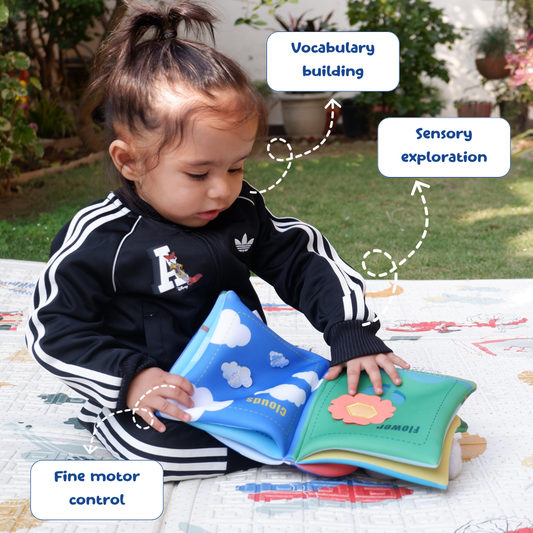 Child playing with a soft book outdoors, with text labels on vocabulary building, sensory exploration, and fine motor control.